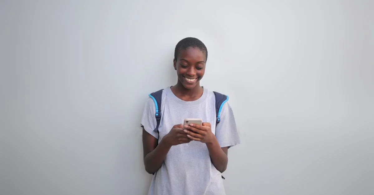 Une jeune femme se tient à l'intérieur utilisant un smartphone, souriant joyeusement.
