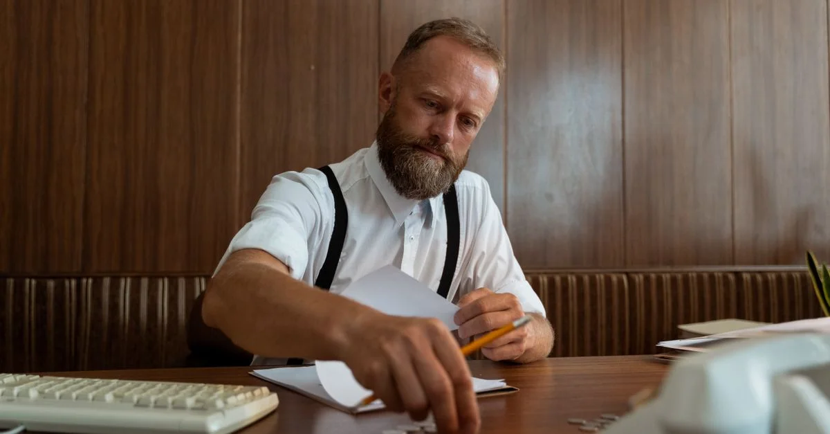 Un homme d'affaires barbu portant des bretelles compte des pièces dans un bureau vintage, incarnant un style de bureau rétro.