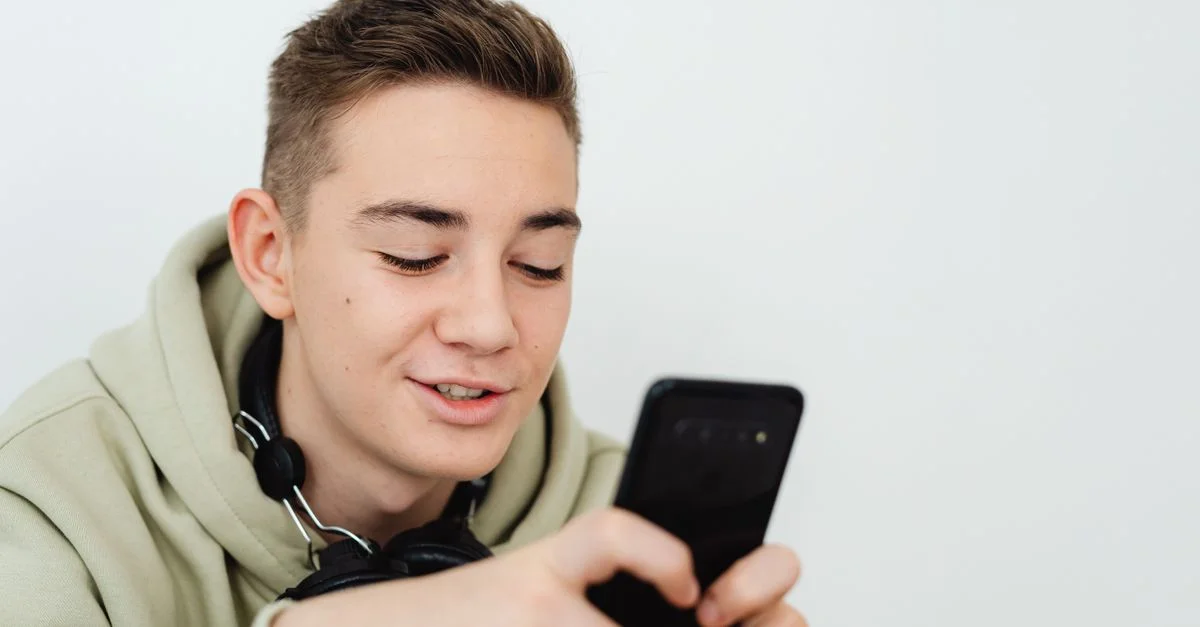 Un jeune homme en sweat à capuche sourit en utilisant son smartphone, des écouteurs autour du cou.